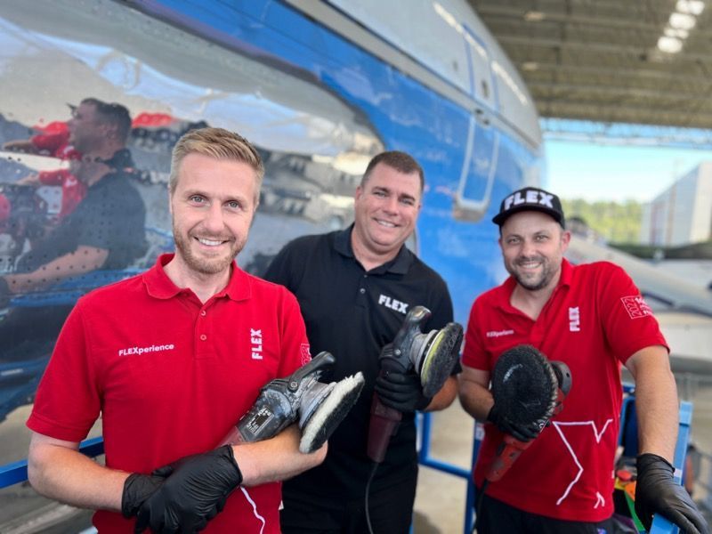 Three men in red FLEX shirts polish a plane; two hold buffers, smiling outdoors.