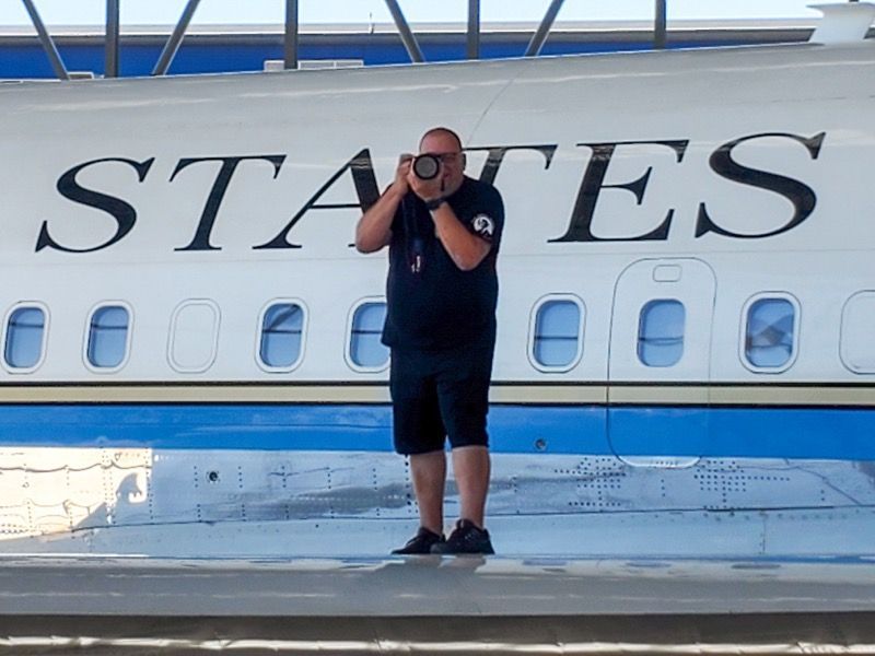 Man taking photo in front of the side of a plane that says “STATES”.