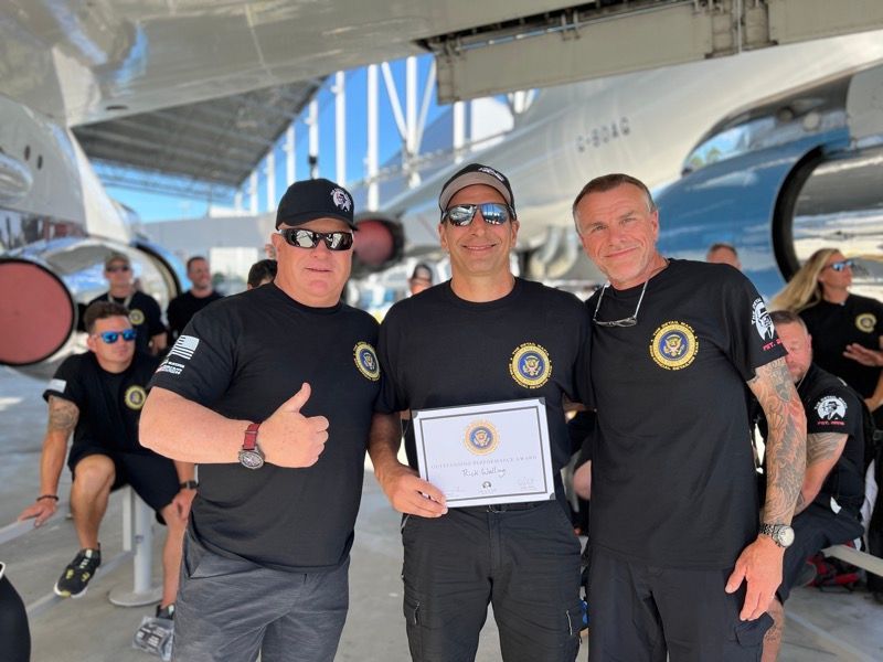 Three men in black shirts pose with a certificate in front of an airplane.