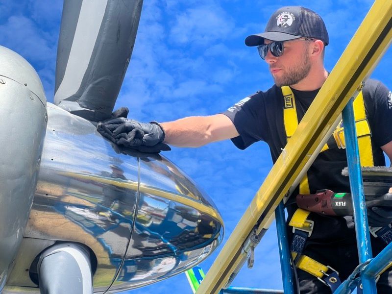 Man polishing aircraft propeller, wearing a cap, sunglasses, and safety harness, against blue sky.