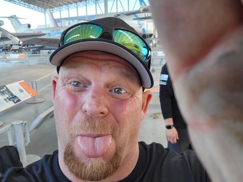 Man with a goatee sticking out his tongue, wearing a hat and sunglasses, at an aviation museum.