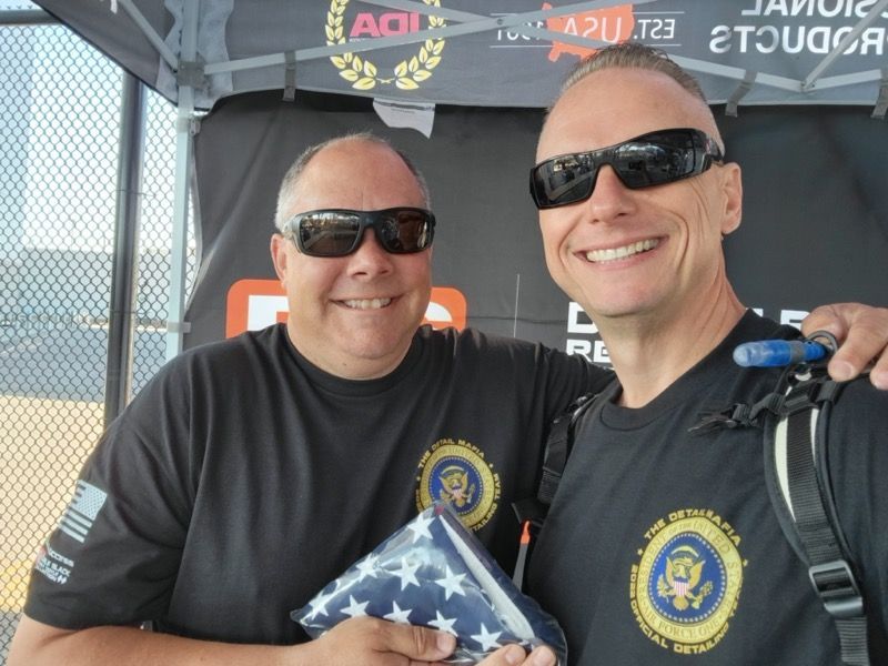 Two men in black shirts with sunglasses smile; one holds a folded American flag.