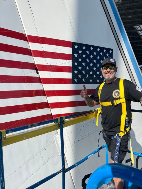 Man giving thumbs-up next to a painted American flag on an aircraft tail.