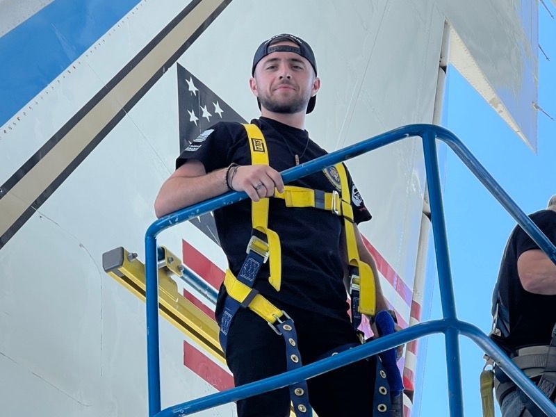 Man in black shirt and hat, safety harness, standing on a platform near a plane with American flag.