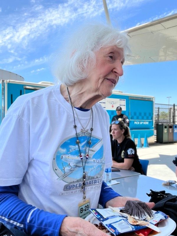 An elderly woman with white hair in a white tee shirt with a blue collar, holding a plate outside, with others nearby.
