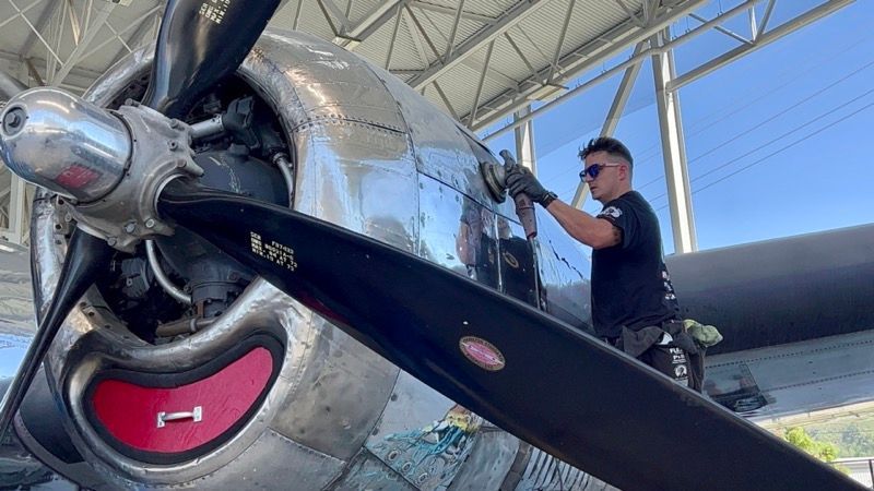 A man polishing the shiny fuselage of a vintage airplane near the propeller.