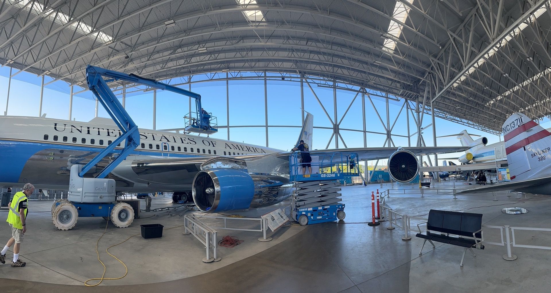 Large airplane in hangar with workers using a lift. Blue and white color scheme, sunny day.
