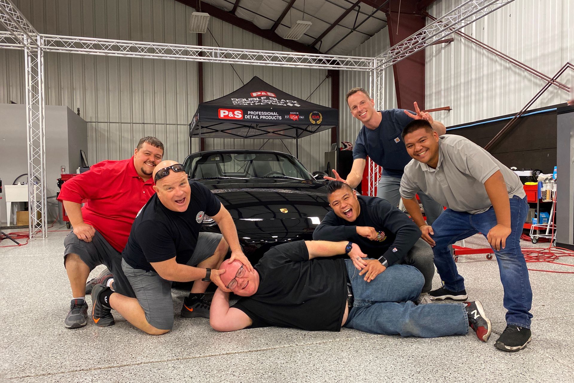 Six men posing playfully with a black car in a garage; men are smiling, with one on the ground.