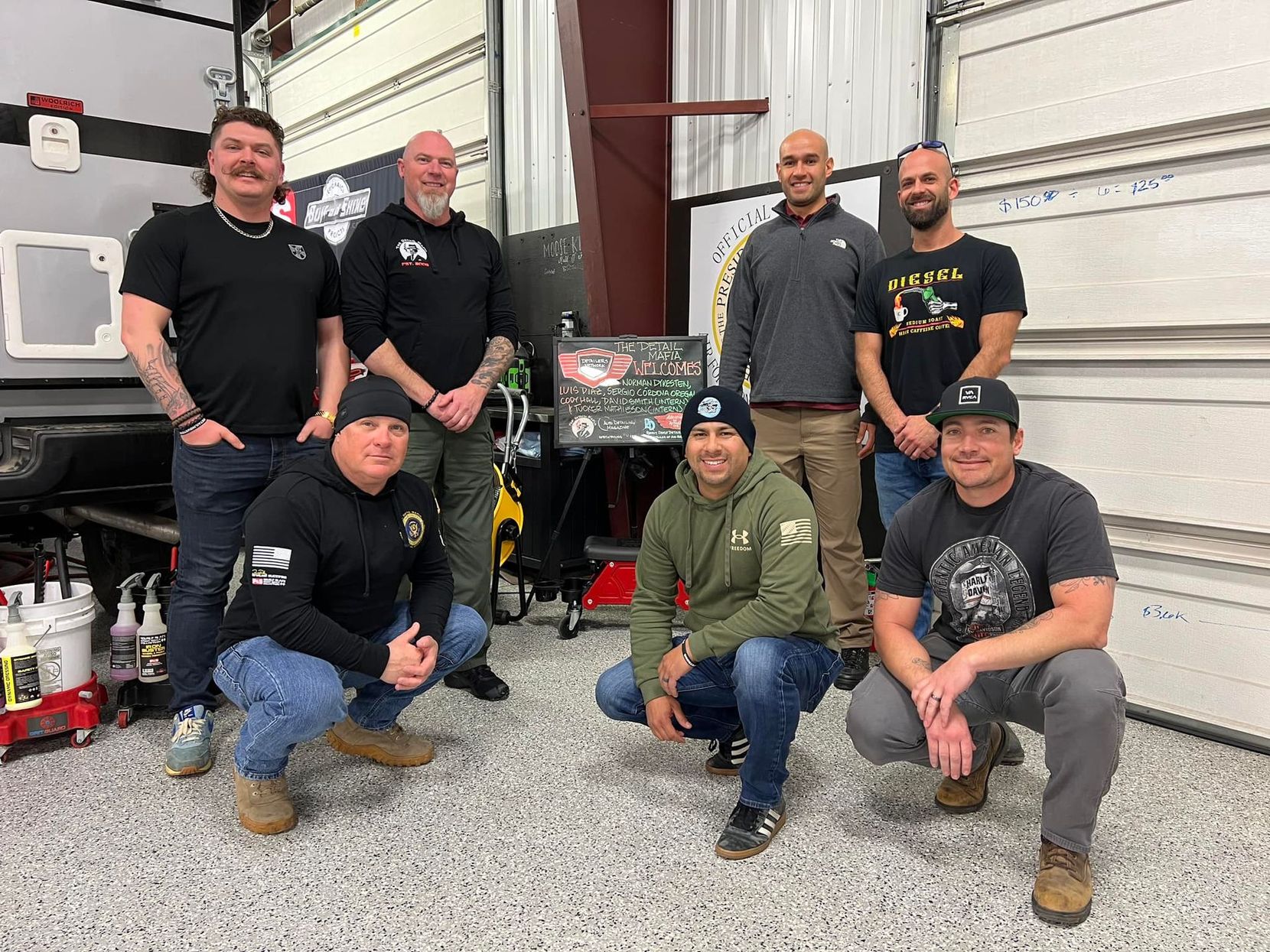 Group of eight men posing together in a garage. Some are kneeling. They are smiling, and the setting appears to be an auto shop.