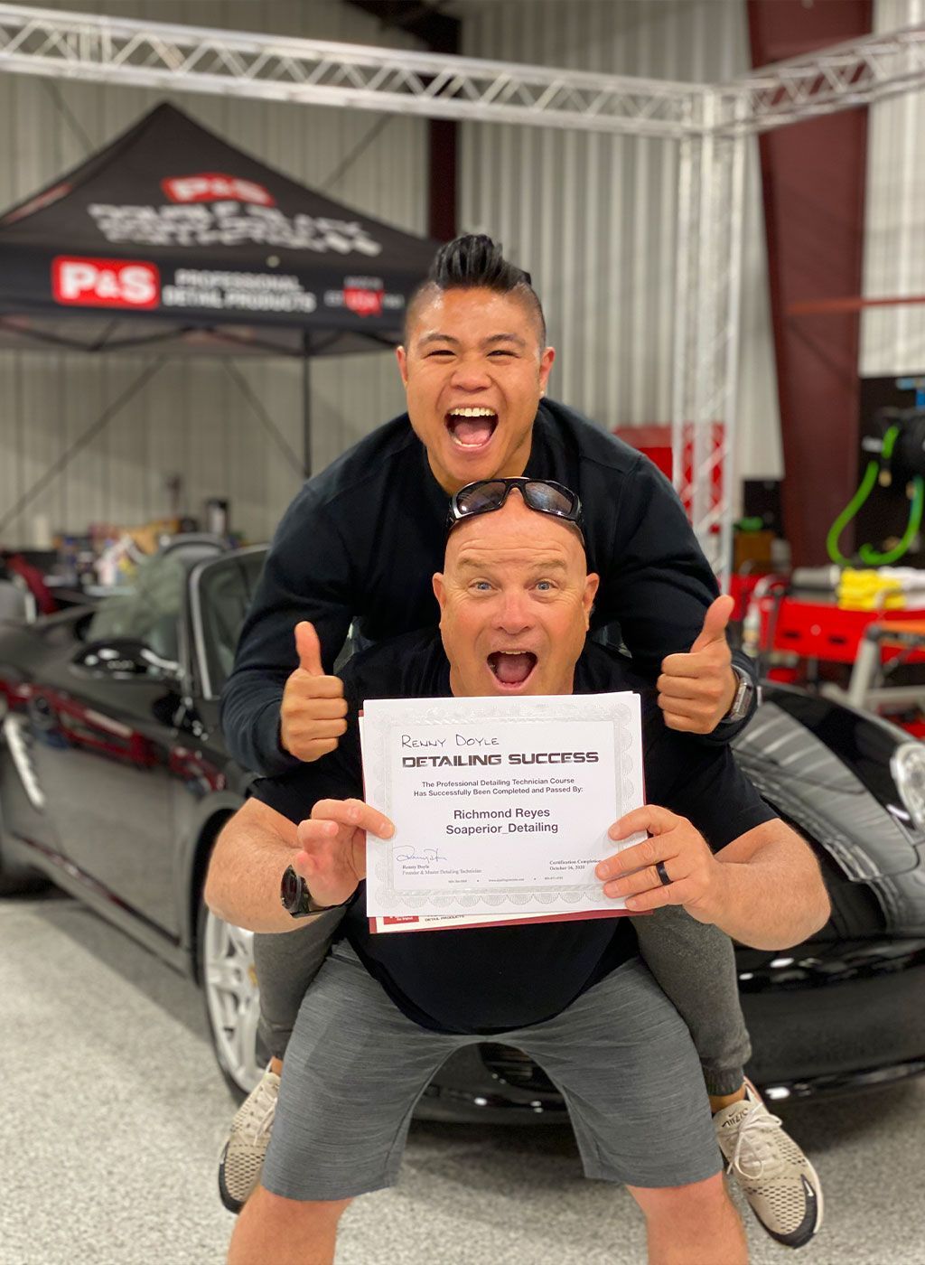 Two men celebrating with thumbs up, one piggybacking the other holding a certificate in front of a black car.
