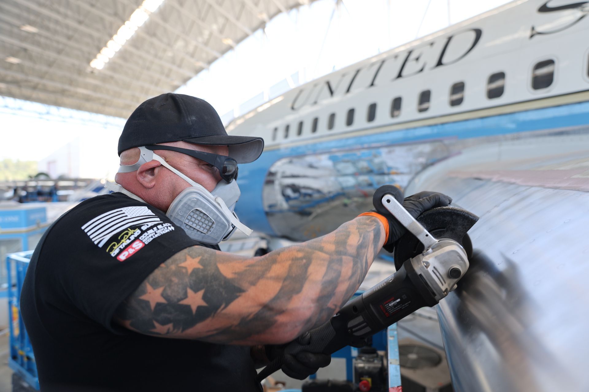 Man with arm tattoos grinds metal on the side of a United Airlines plane.