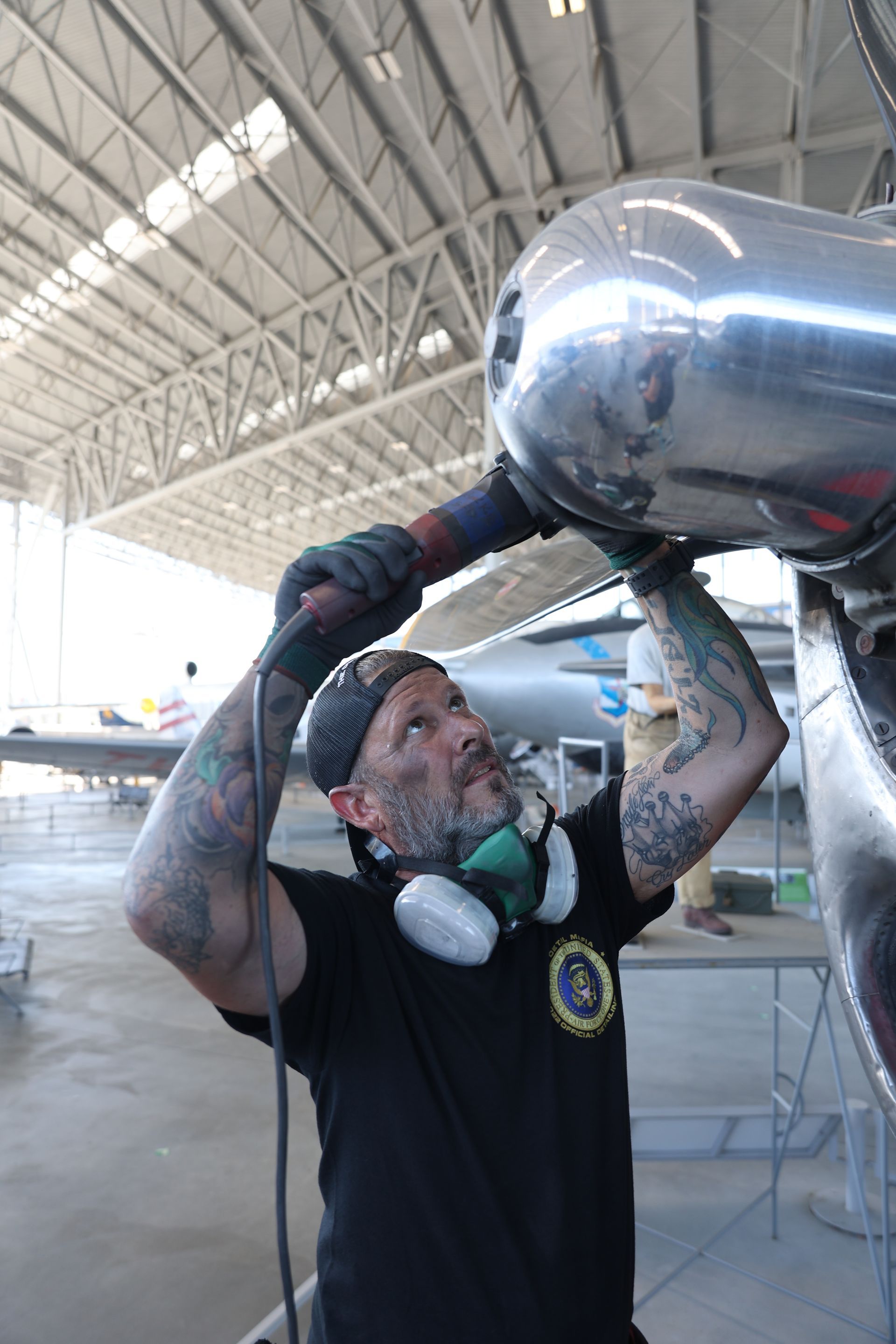 Man with tattoos polishes airplane part in hangar; wears respirator, black shirt, and beanie.