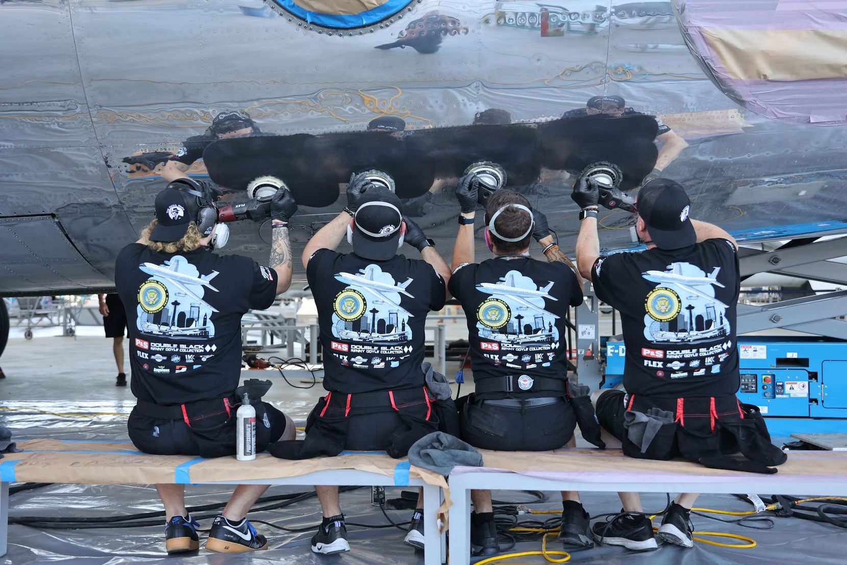Four people wearing black shirts work on an aircraft, using tools to repair it.