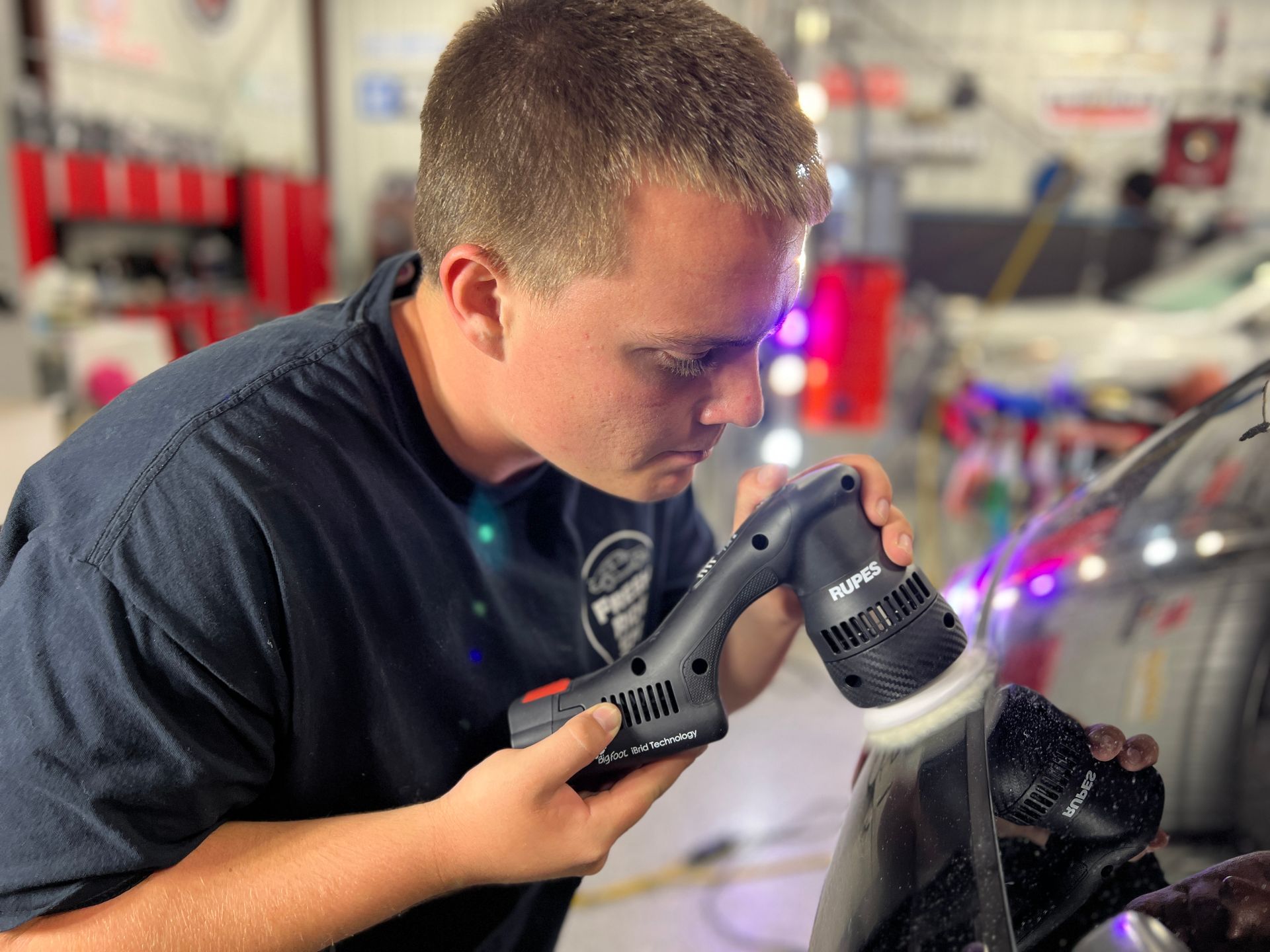 Mechanic polishes a car window with a handheld buffer in a repair shop.