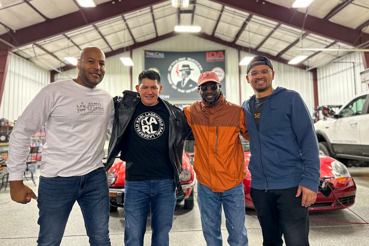 Four men standing in a garage with arms around each other and a red car in the background.