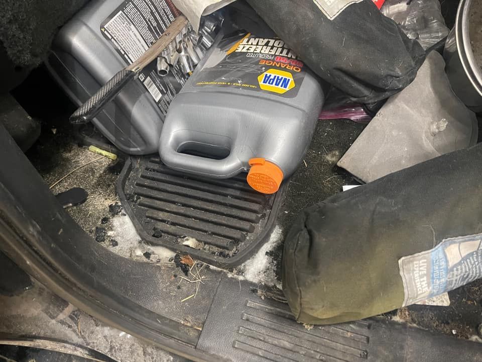 Car interior filled with clutter: antifreeze, black bags, and a floor mat.