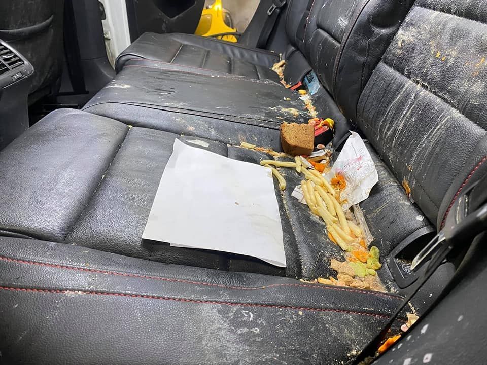 Rear car seat covered in food waste, including fries, sandwich, and paper. Black leather with red stitching.