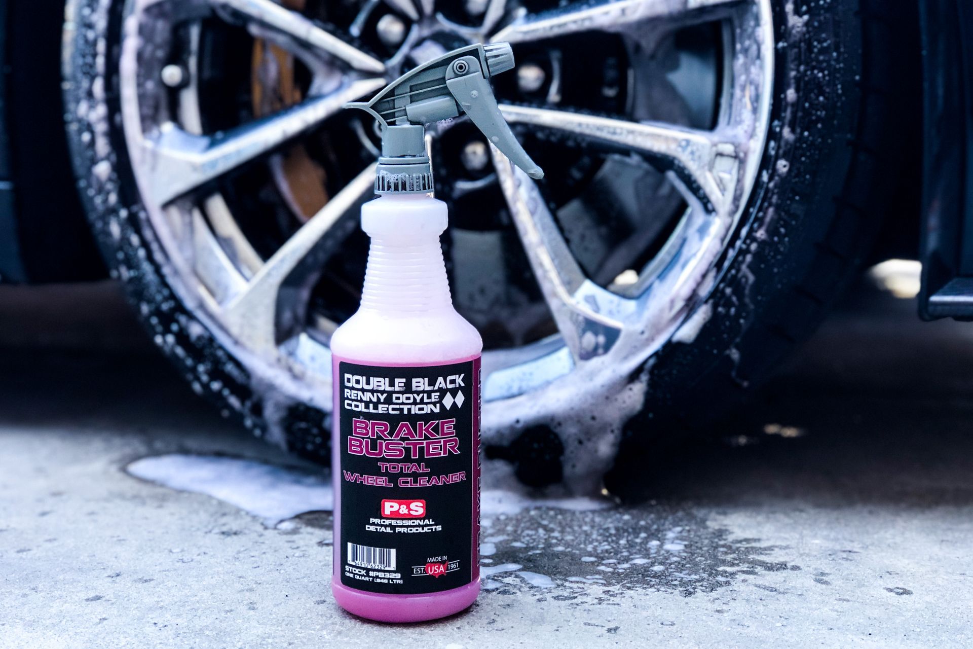 Pink spray bottle in front of a car wheel, covered in foam, for cleaning tires.