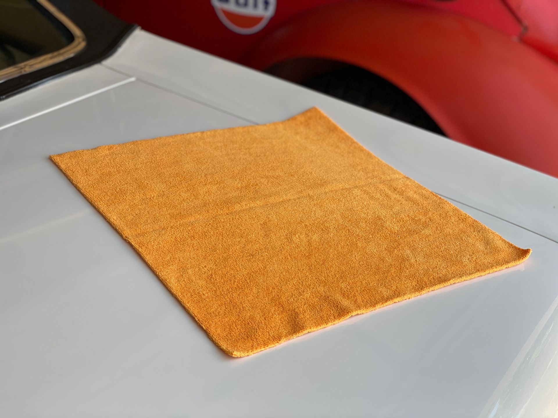 Orange microfiber cloth on the white hood of a car. A red car is in the background.