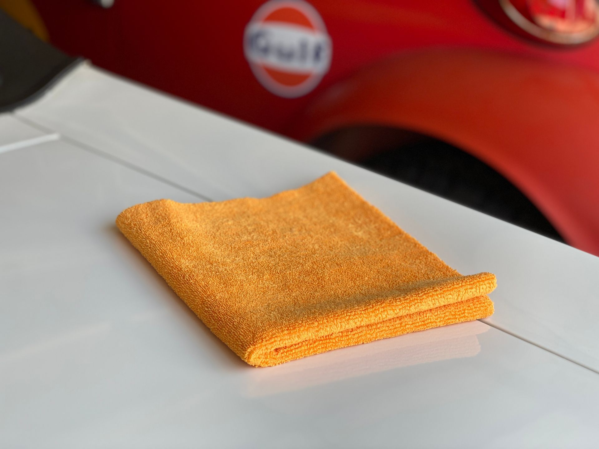 Orange microfiber towel folded on a white car surface, red car with a Gulf sticker in the background.