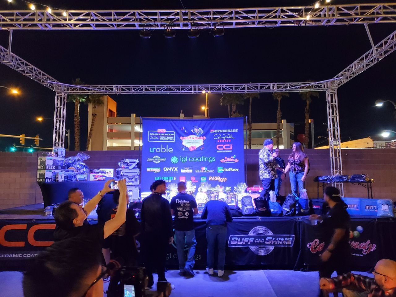 People on stage at an outdoor event with a banner of sponsors. Audience members take photos.