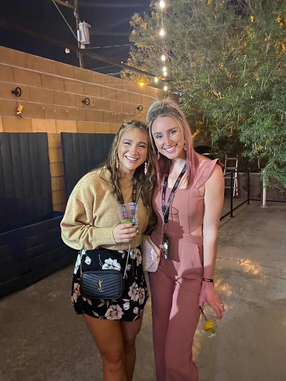 Two women smiling, posing outdoors at night. One wears a pink jumpsuit, the other a tan sweater and floral skirt.
