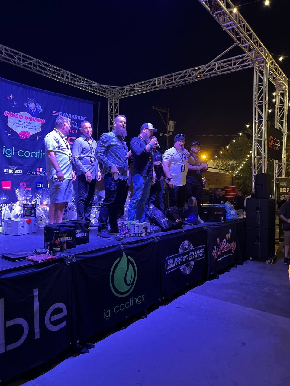 Group of men on stage at night, one speaking into a microphone, event banner in background.