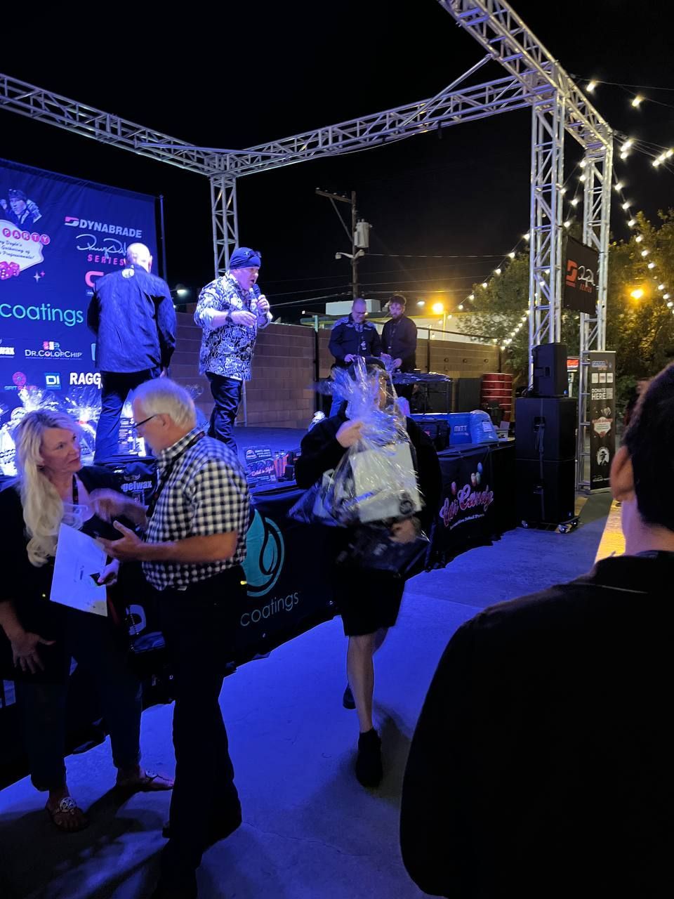People at an outdoor stage at night. One man speaks into a microphone. Others stand near the stage.