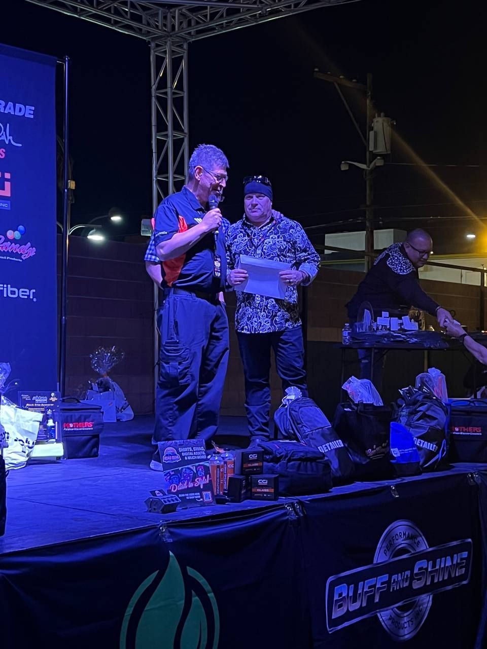 Two men on a stage, one speaking into a microphone, the other holding papers. Dark setting, event.