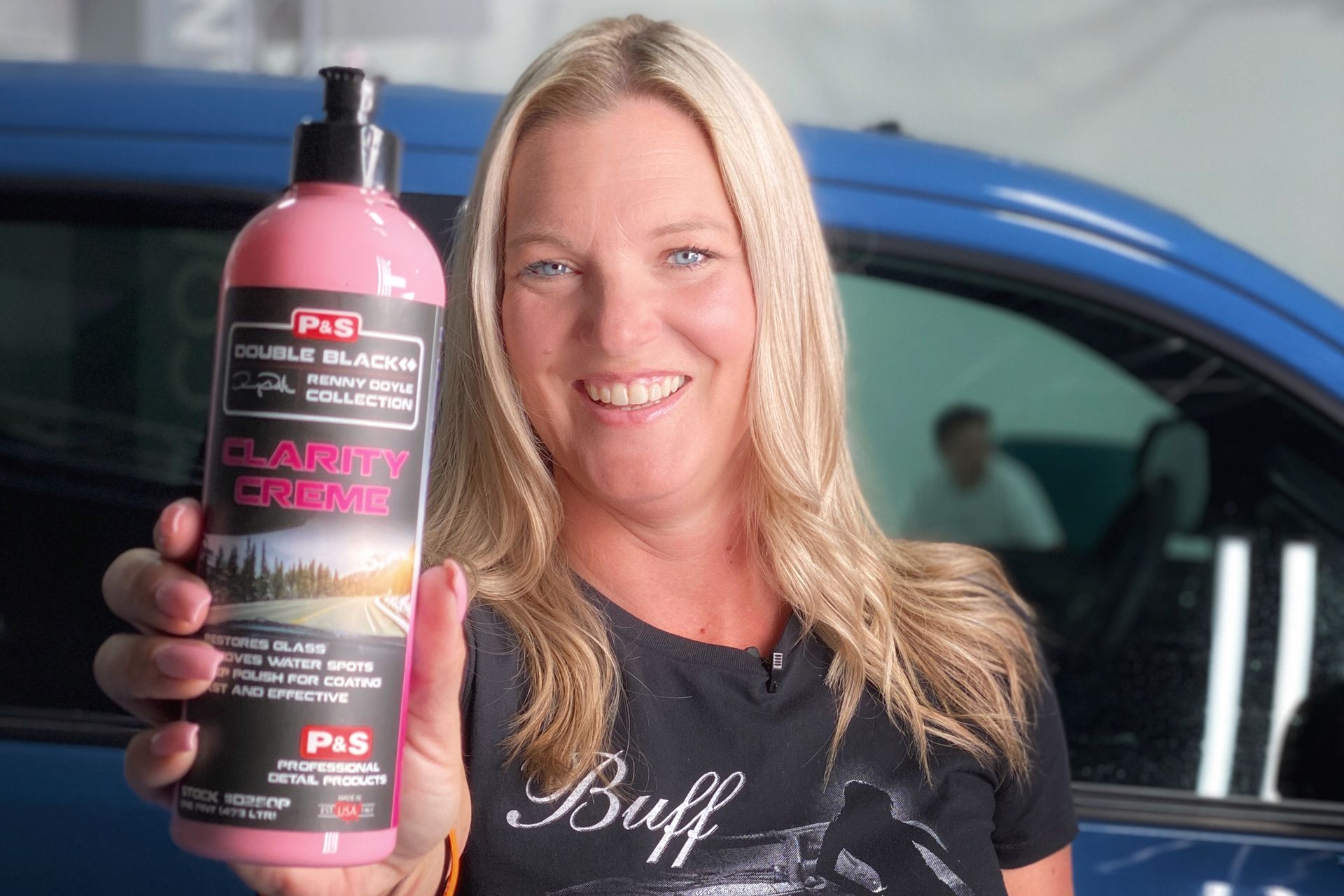Woman holding pink car detailing product, smiling. Blue car in background.