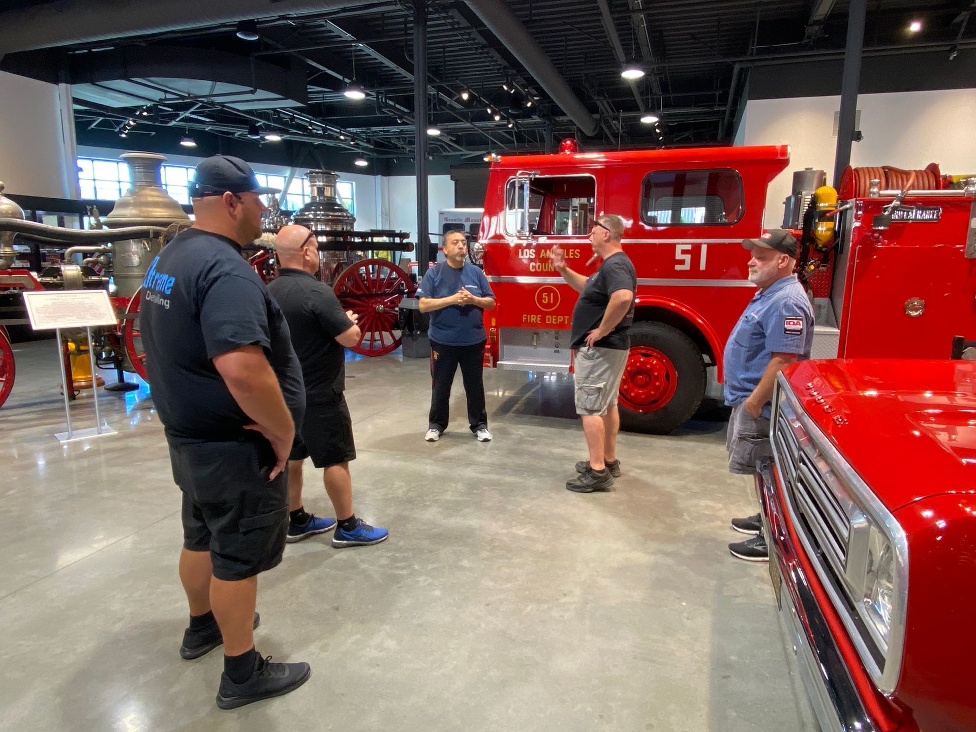 Men in a fire truck museum; one speaks as others listen near red trucks.