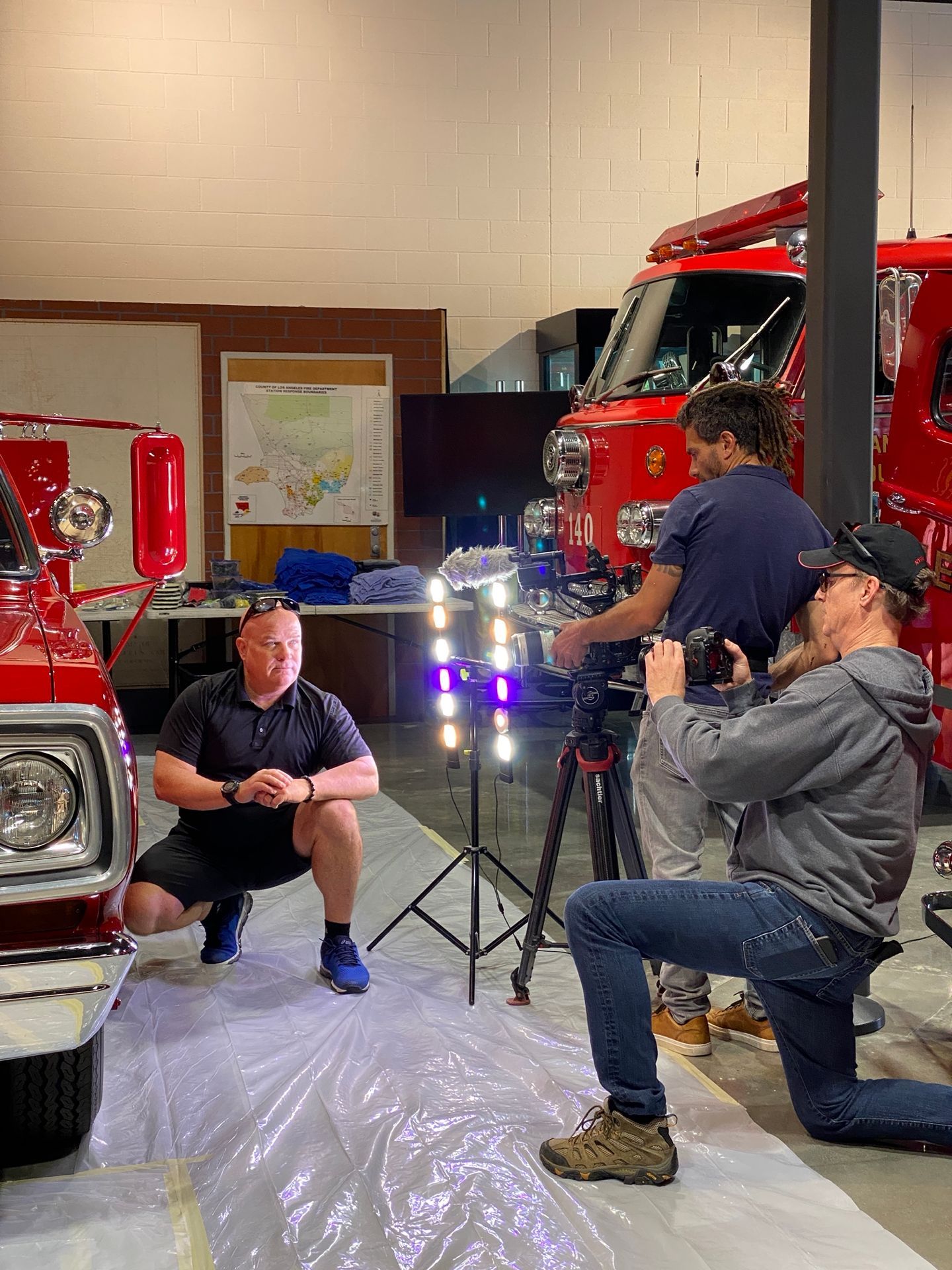 Man squats in front of a fire truck, being filmed by two people in a fire station.
