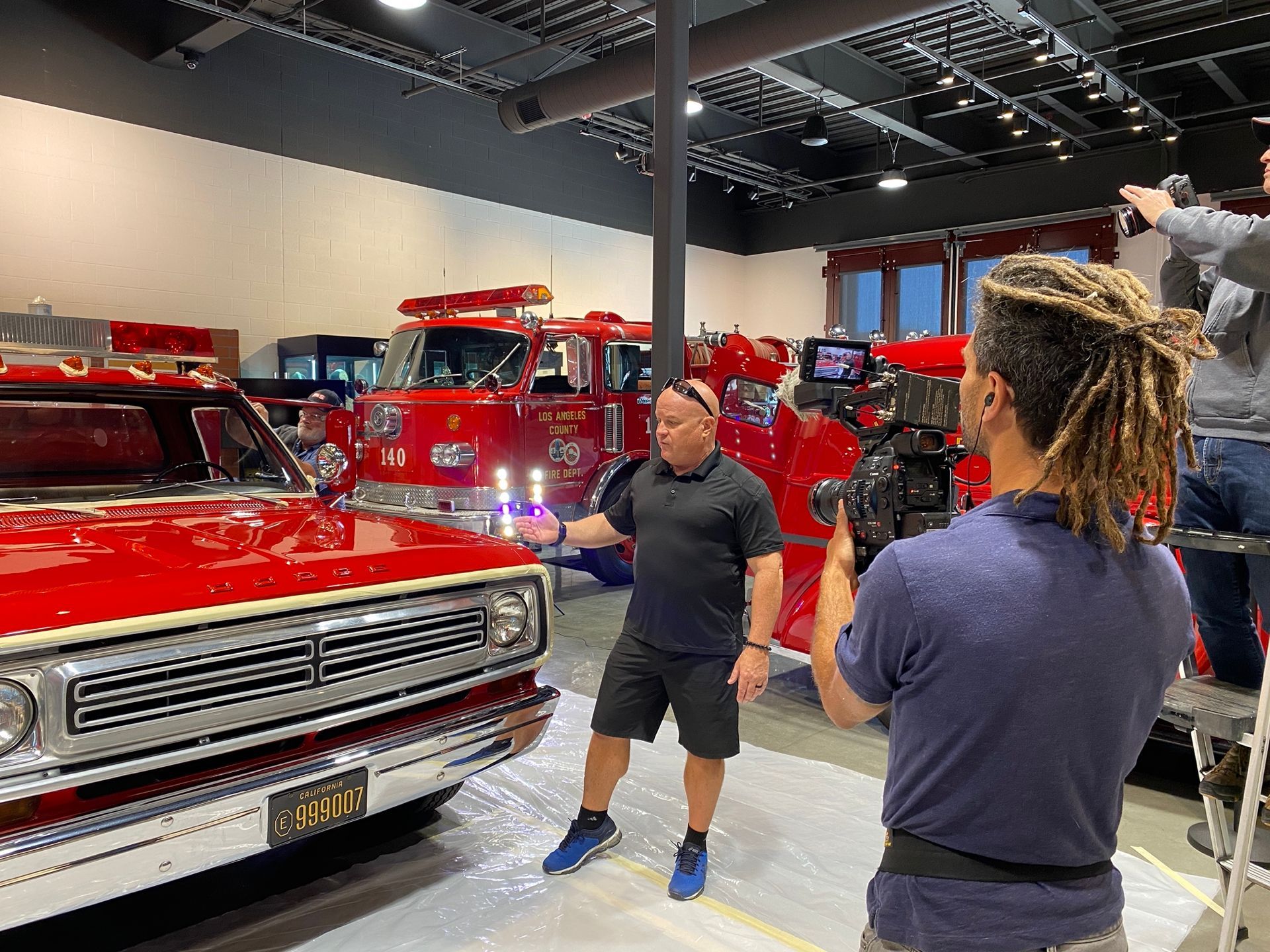 Man in black shirt being filmed in front of a red fire truck and classic red vehicle.