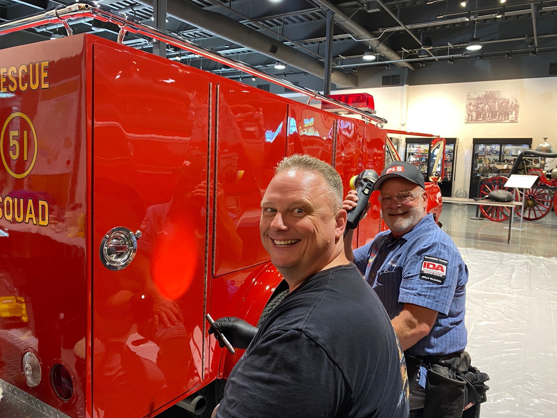 Two men smiling while working on a bright red fire truck in an indoor setting.