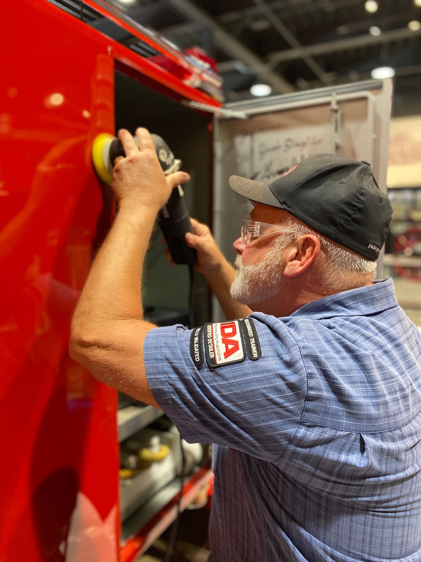 Man polishing a red vehicle with a power polisher. He wears a hat and blue shirt.