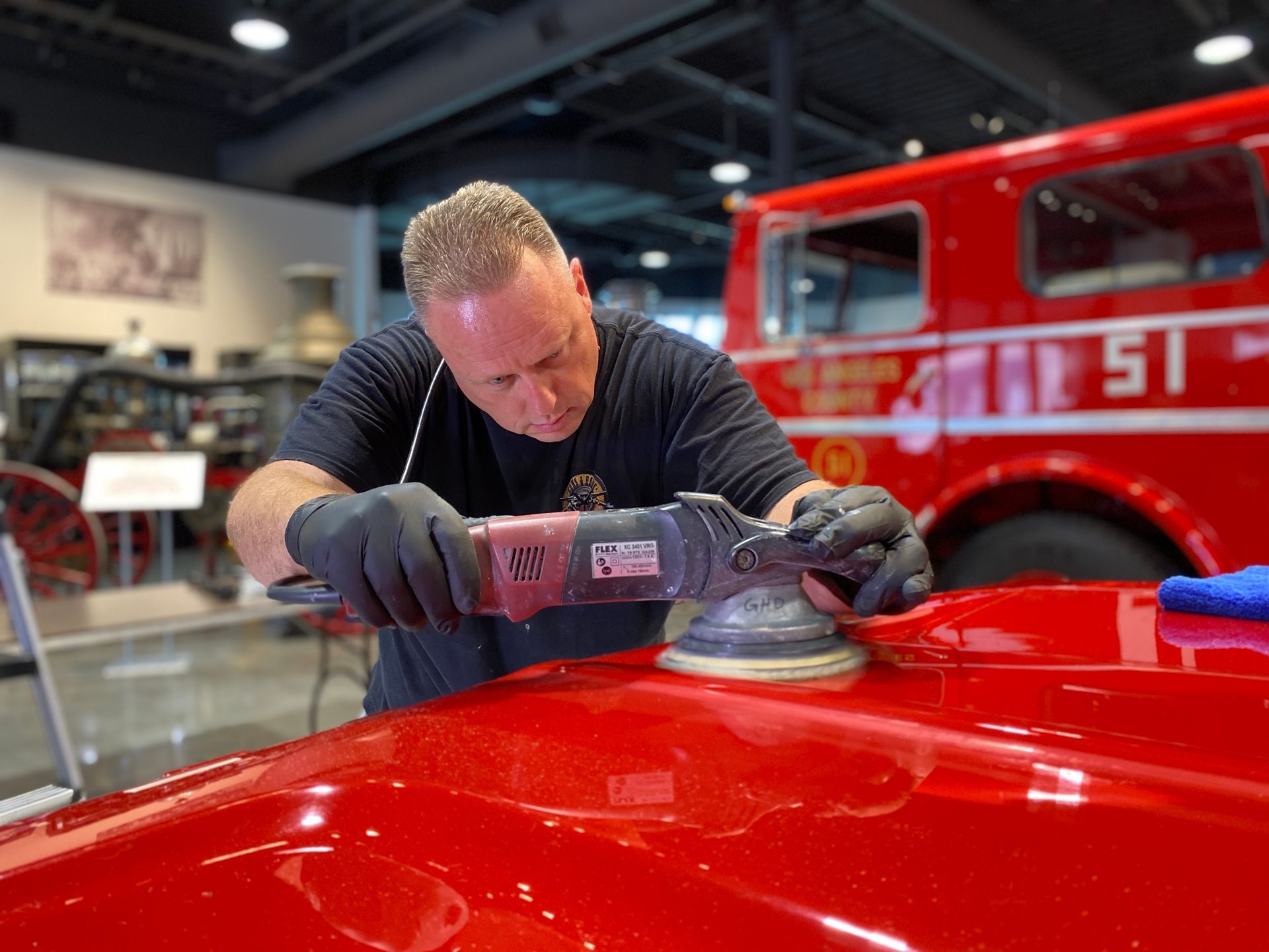 Man polishing a red fire truck with a power buffer. Wearing black gloves, inside a museum-like space.