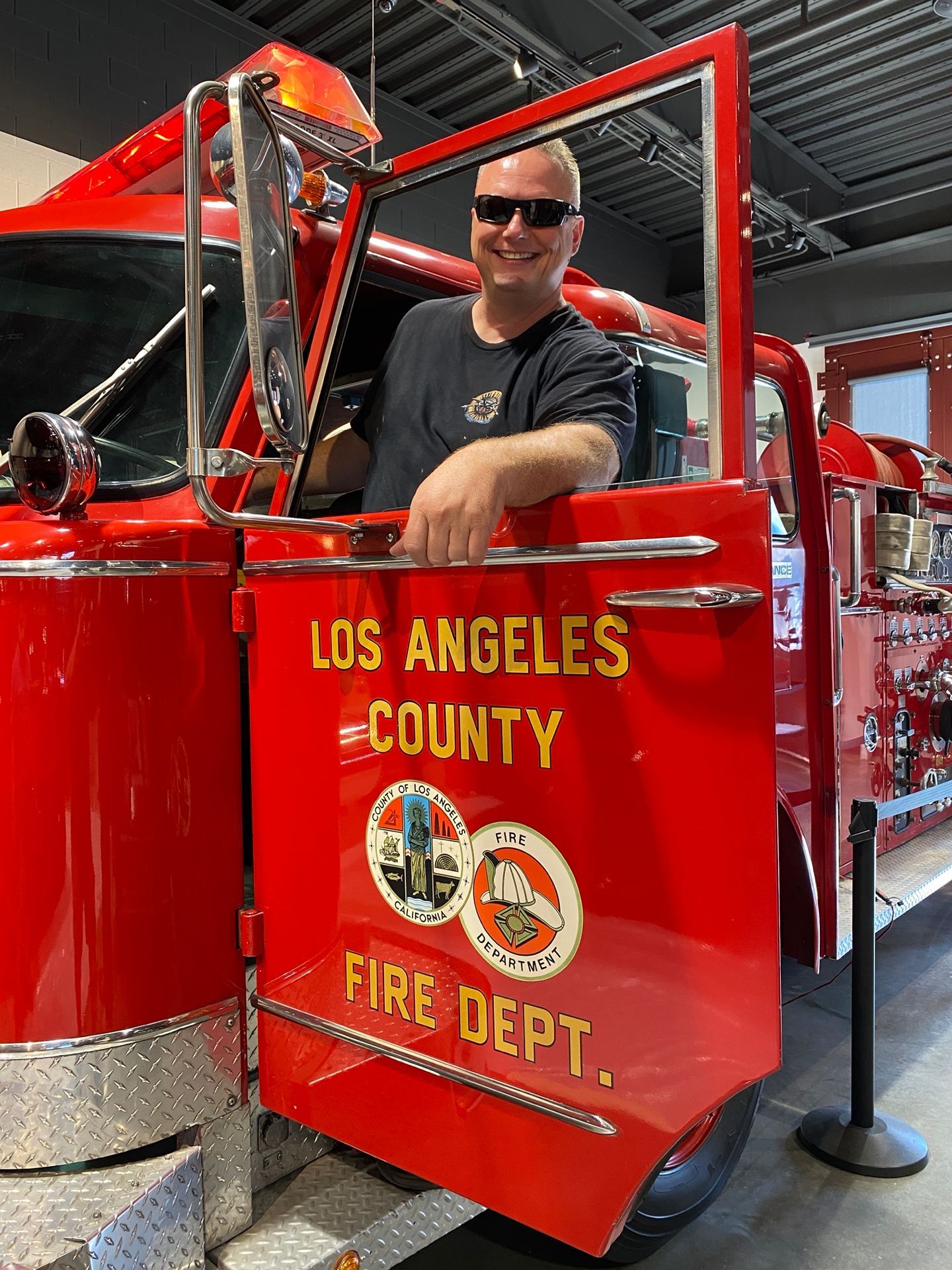 Man smiles, leaning out of a vintage Los Angeles County Fire Dept. fire truck.