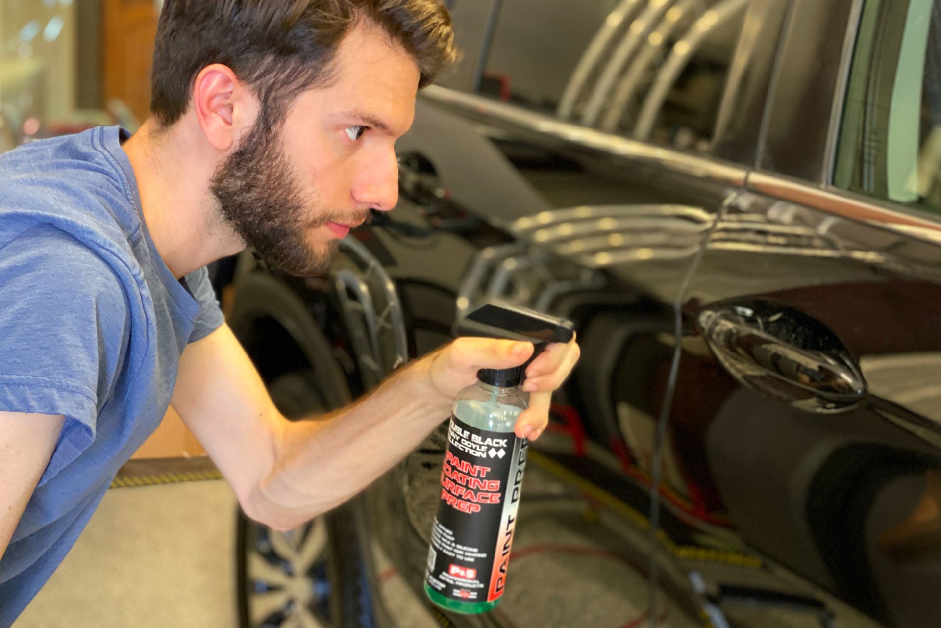 Man sprays cleaning product on a black car door, likely detailing in a garage.