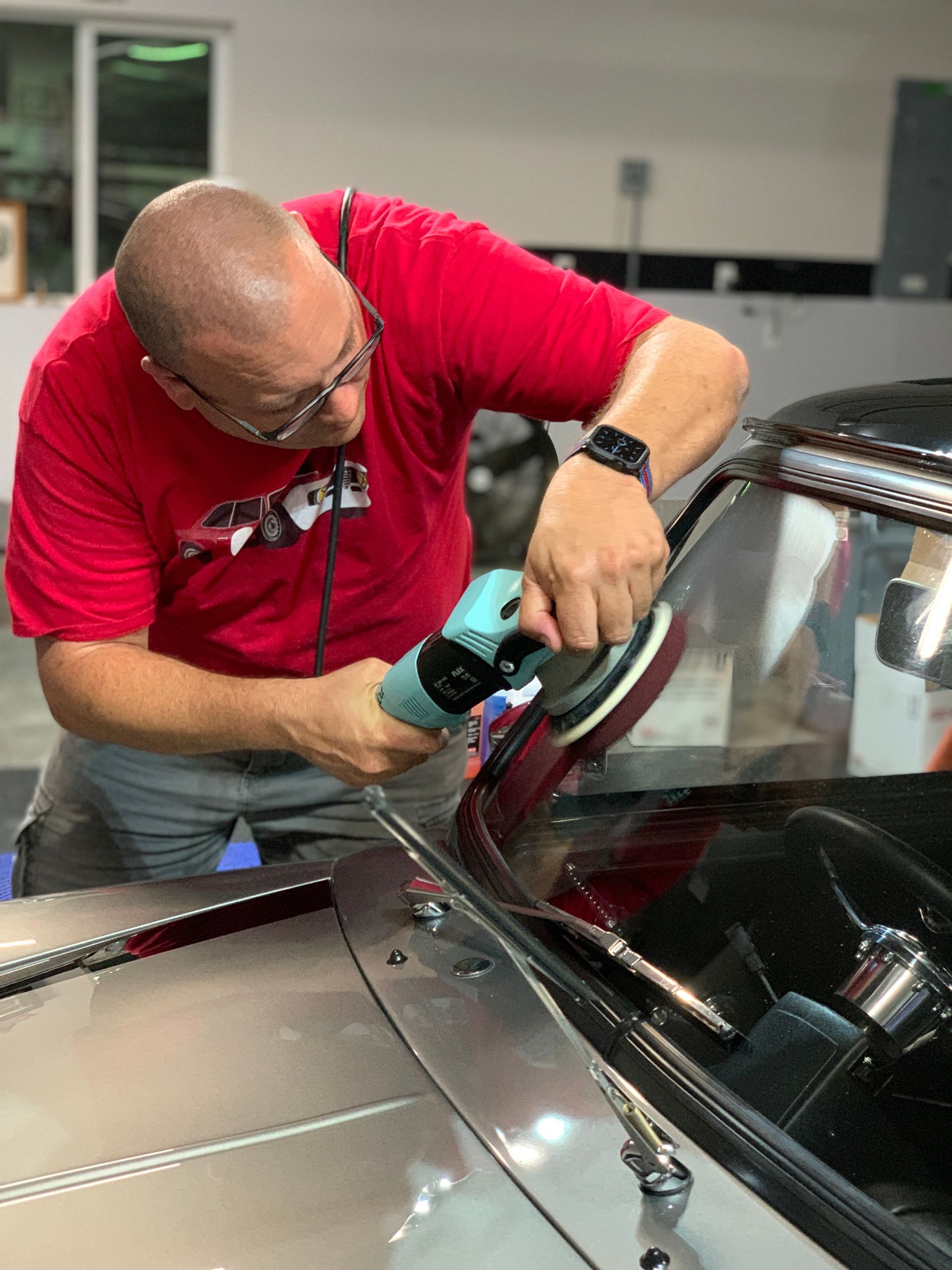 A man in a red shirt polishing a car windshield with a buffing machine.