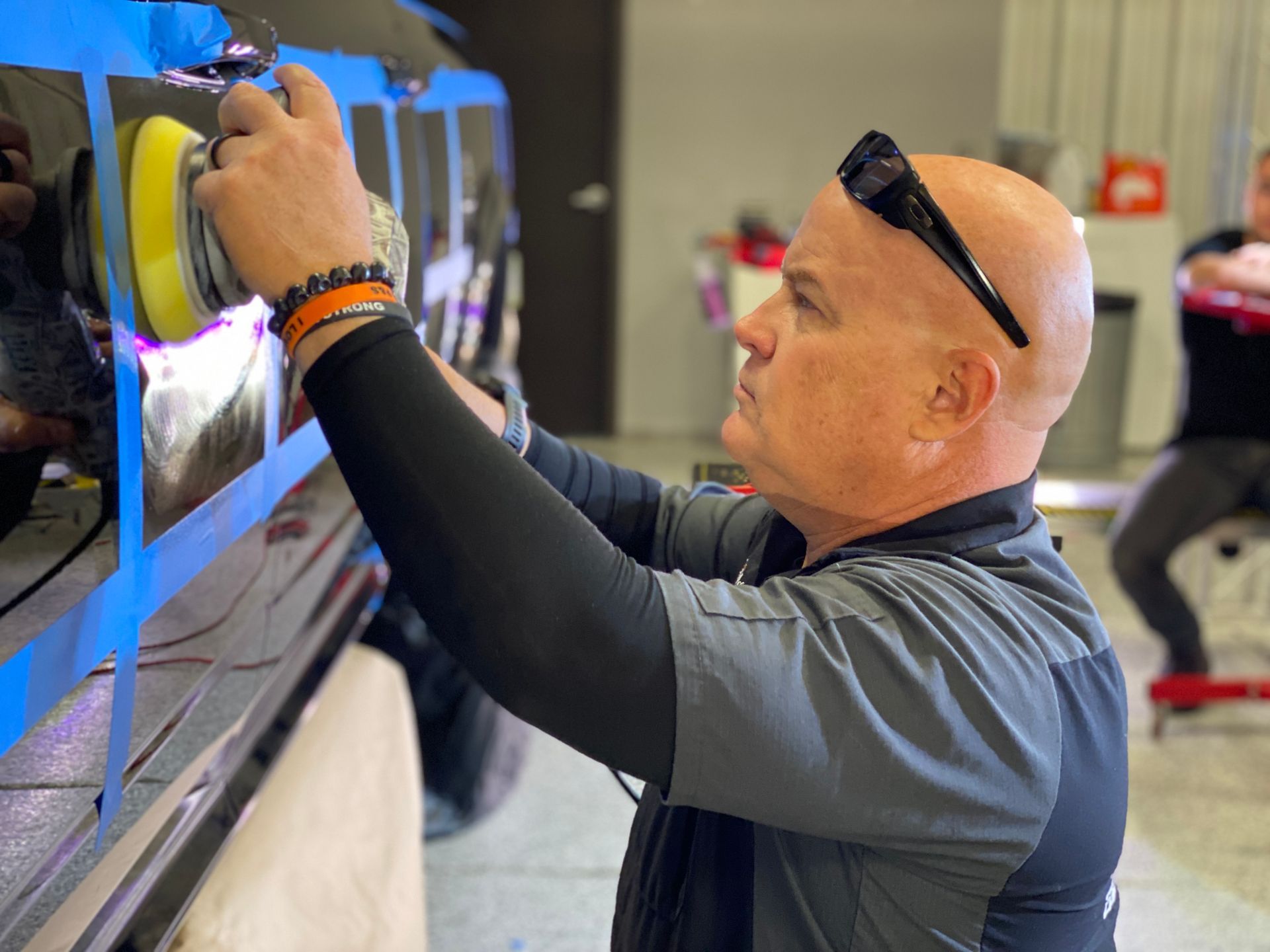 Man using a polisher on a car. He's wearing sunglasses and a black sleeve. Shop setting with another person.
