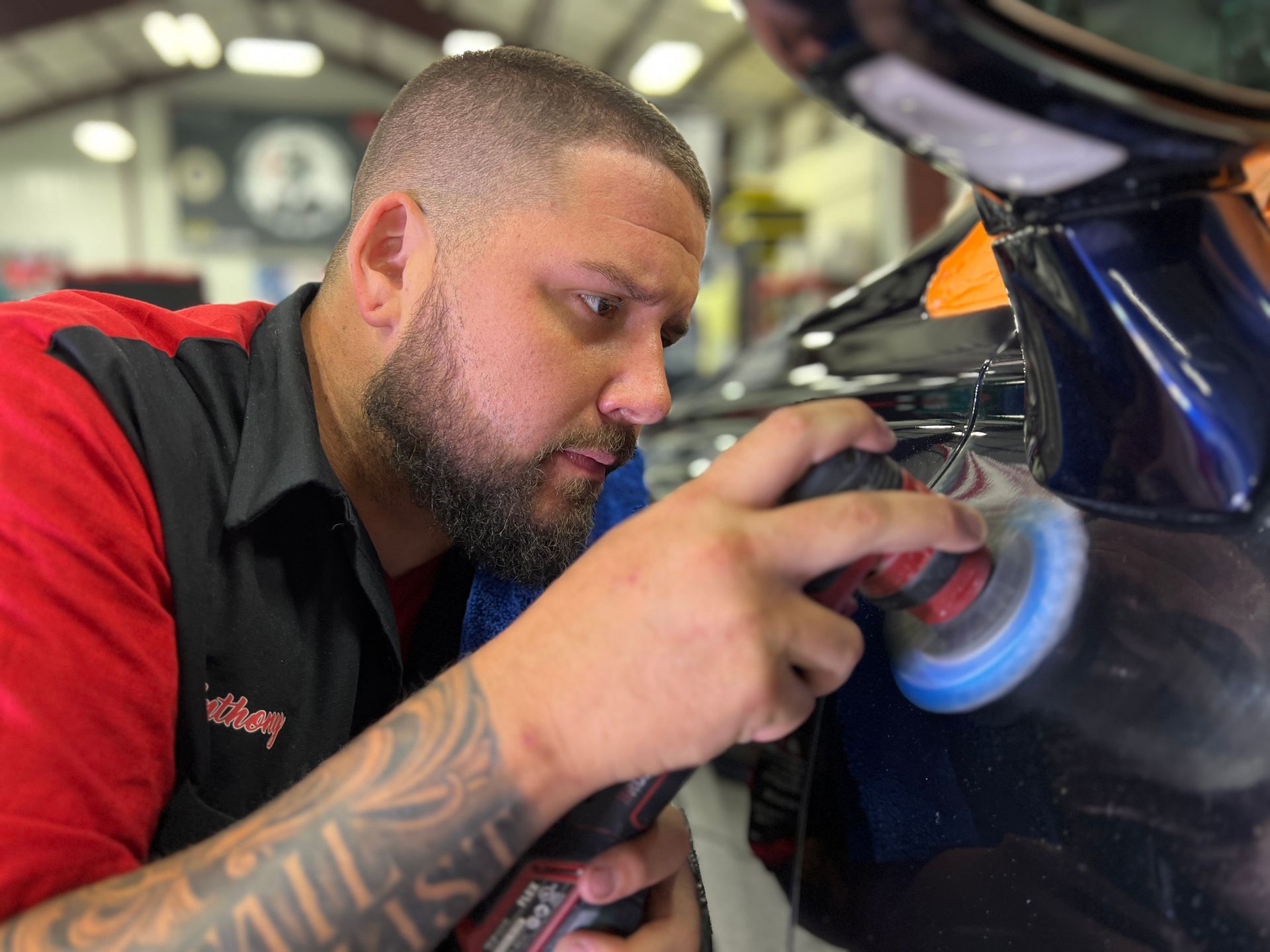 A man polishes a car. He wears a red shirt, has a beard and tattoos. Indoors, focusing on the vehicle.