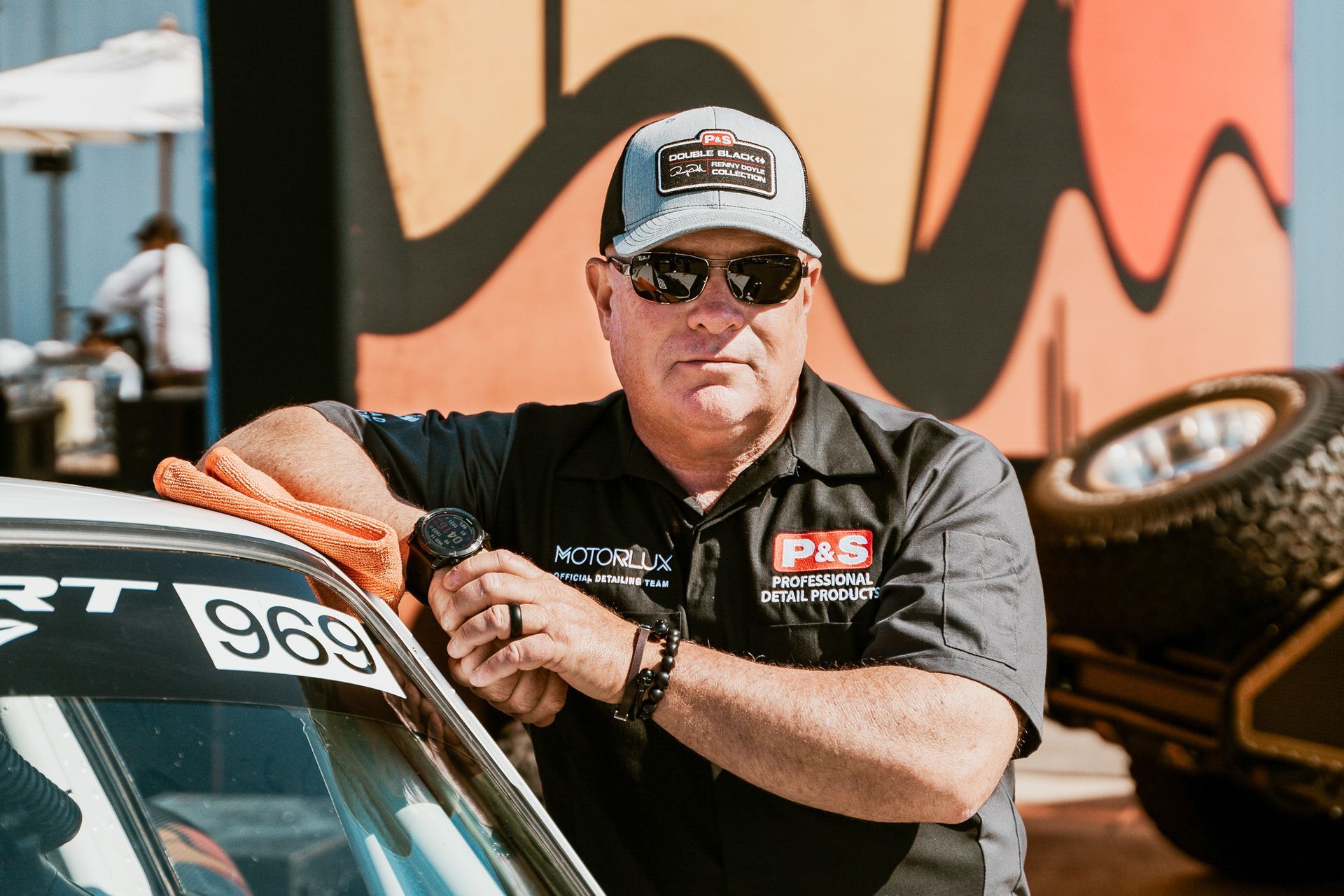 Man in hat and sunglasses, leaning on race car, holding watch, with a colorful background.