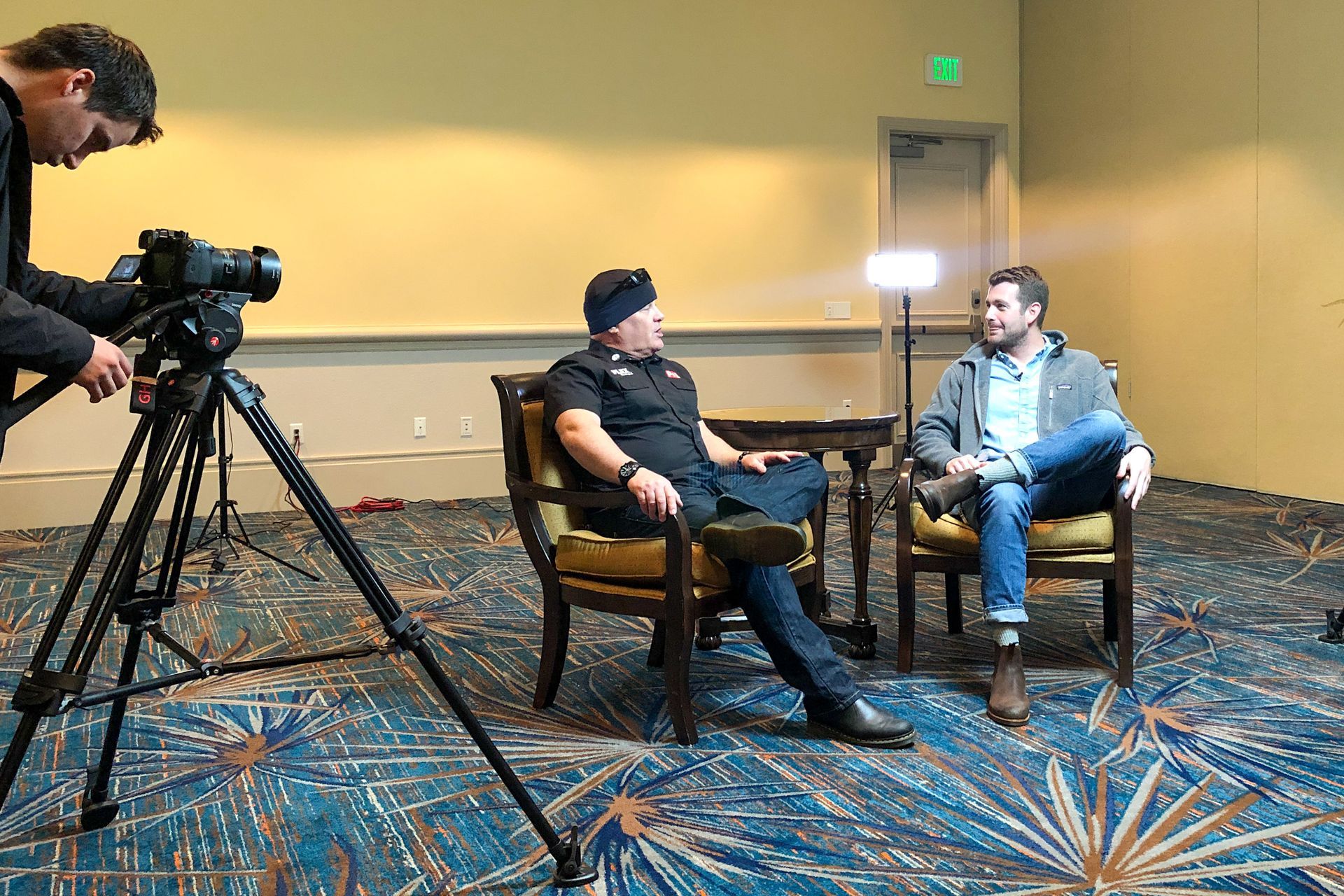 A film crew interviews two men sitting in armchairs in a room with blue patterned carpet.