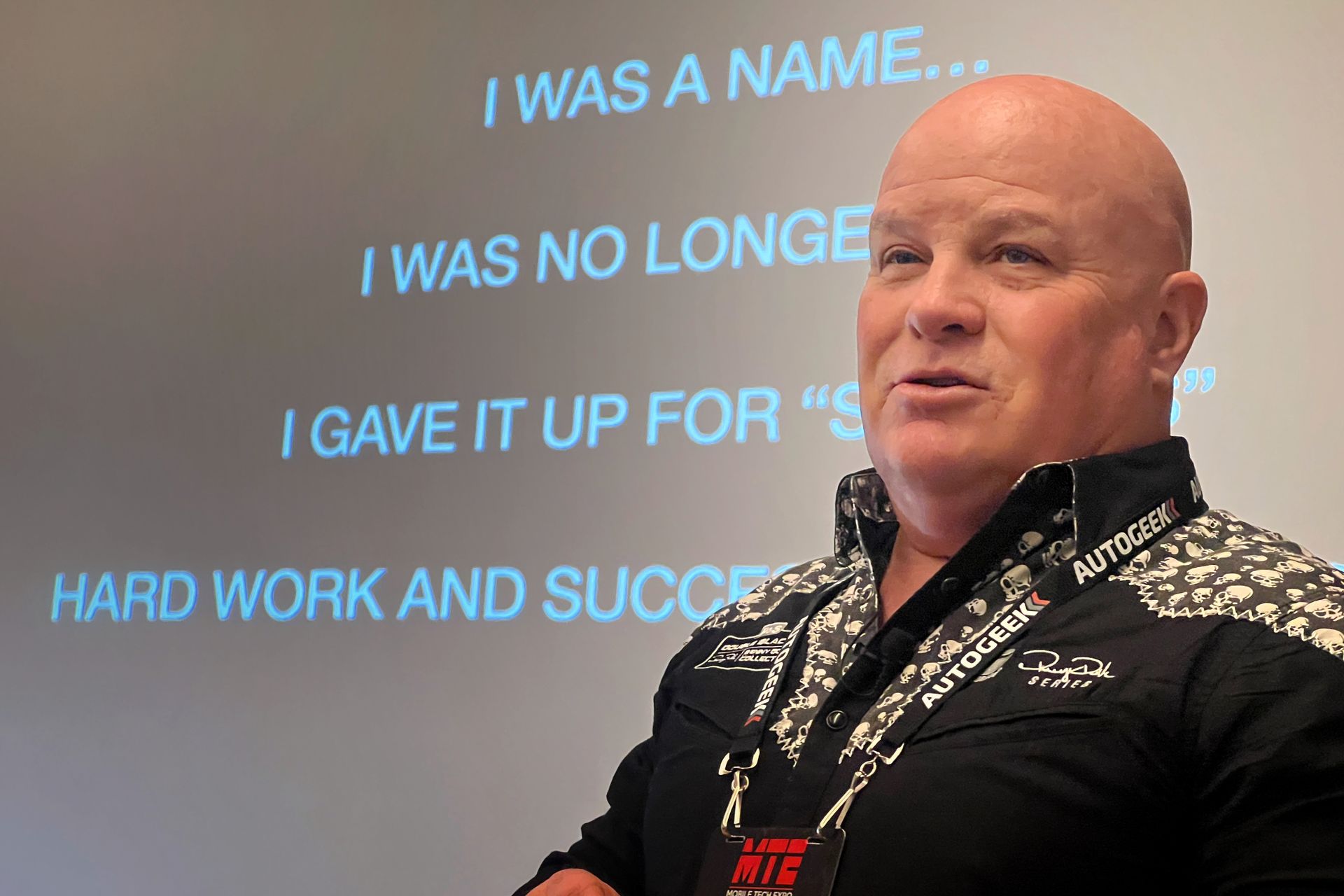 Bald man giving a presentation, words on the screen. Wearing black and white patterned shirt.