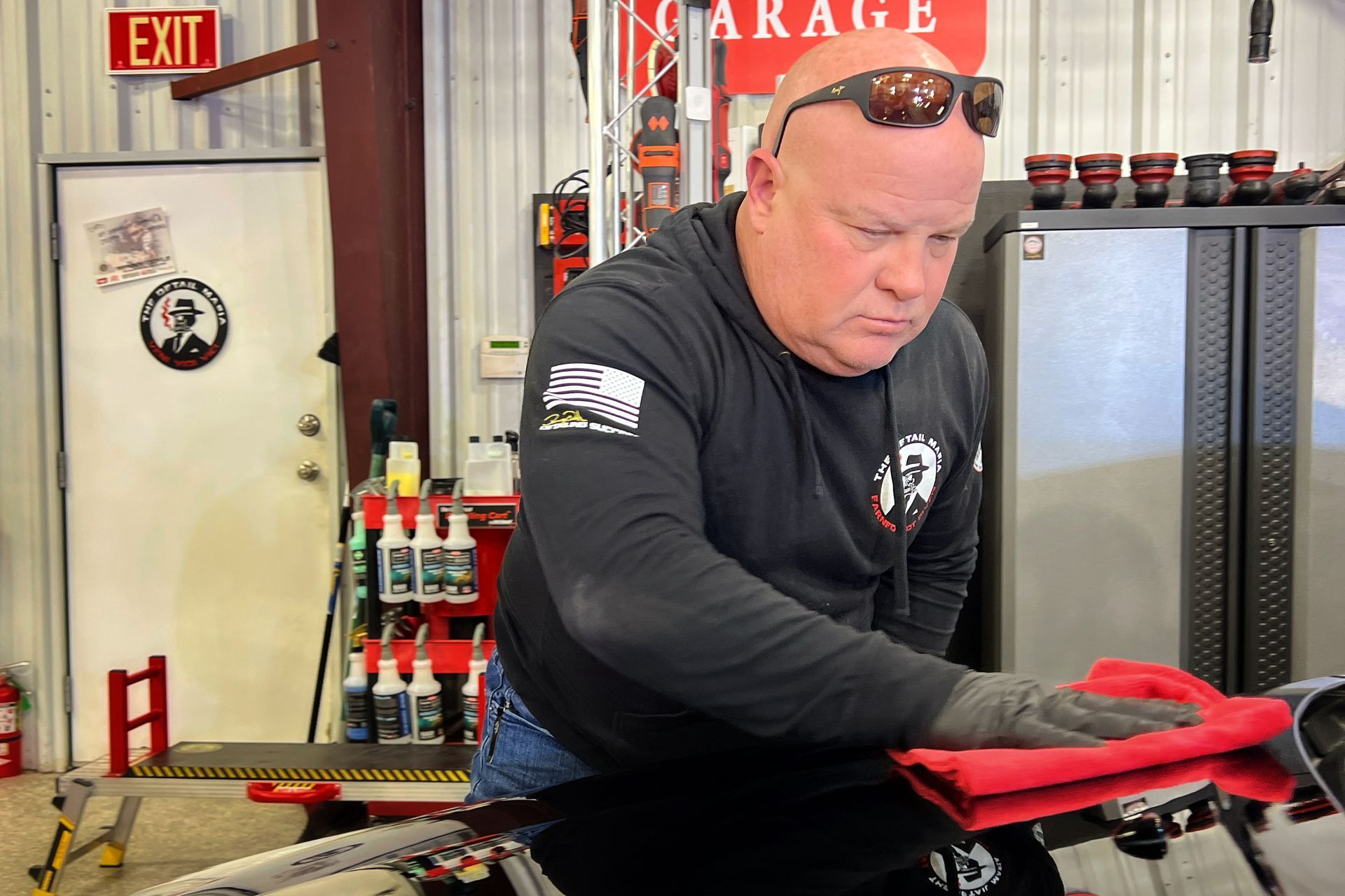 Man in a garage, wearing sunglasses and a black hoodie, polishing a car with a red cloth.