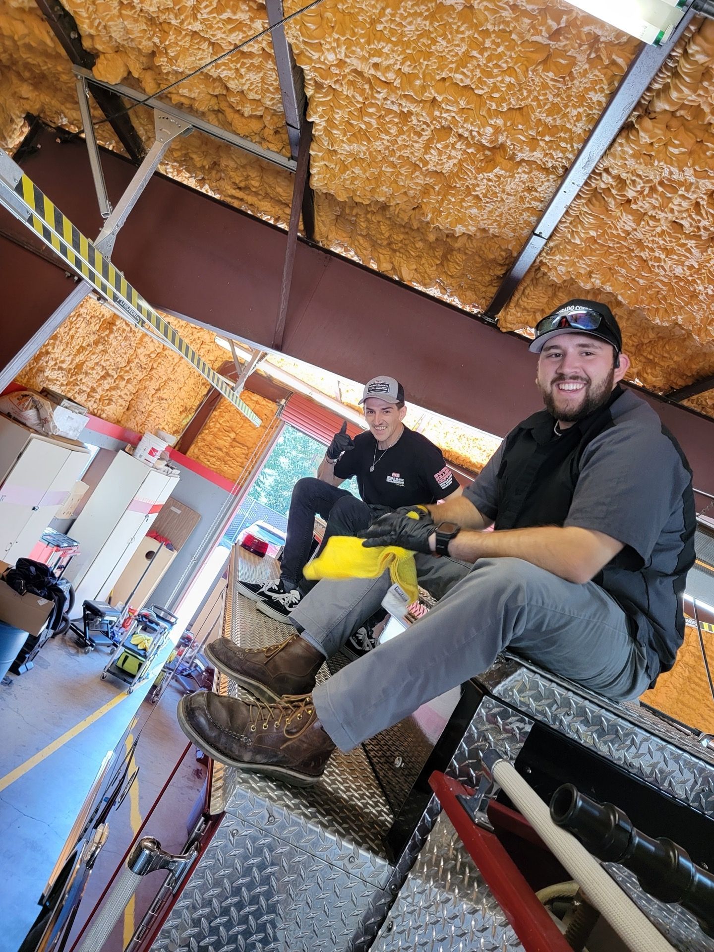 Two men smiling, sitting on a fire truck, inside a garage-like building. One man has a beard and sunglasses.