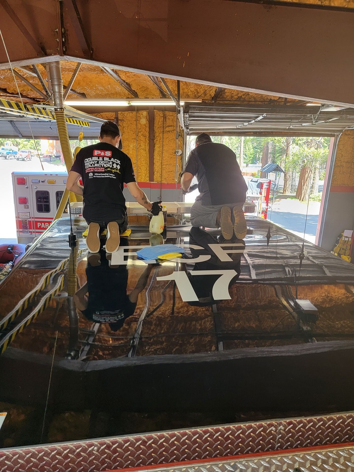 Two men applying decals to a black fire truck roof, under a shelter.