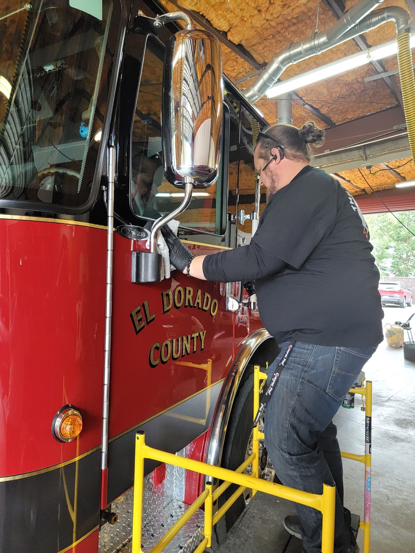 Man on lift working on side mirror of a red and black El Dorado County fire truck in a garage.