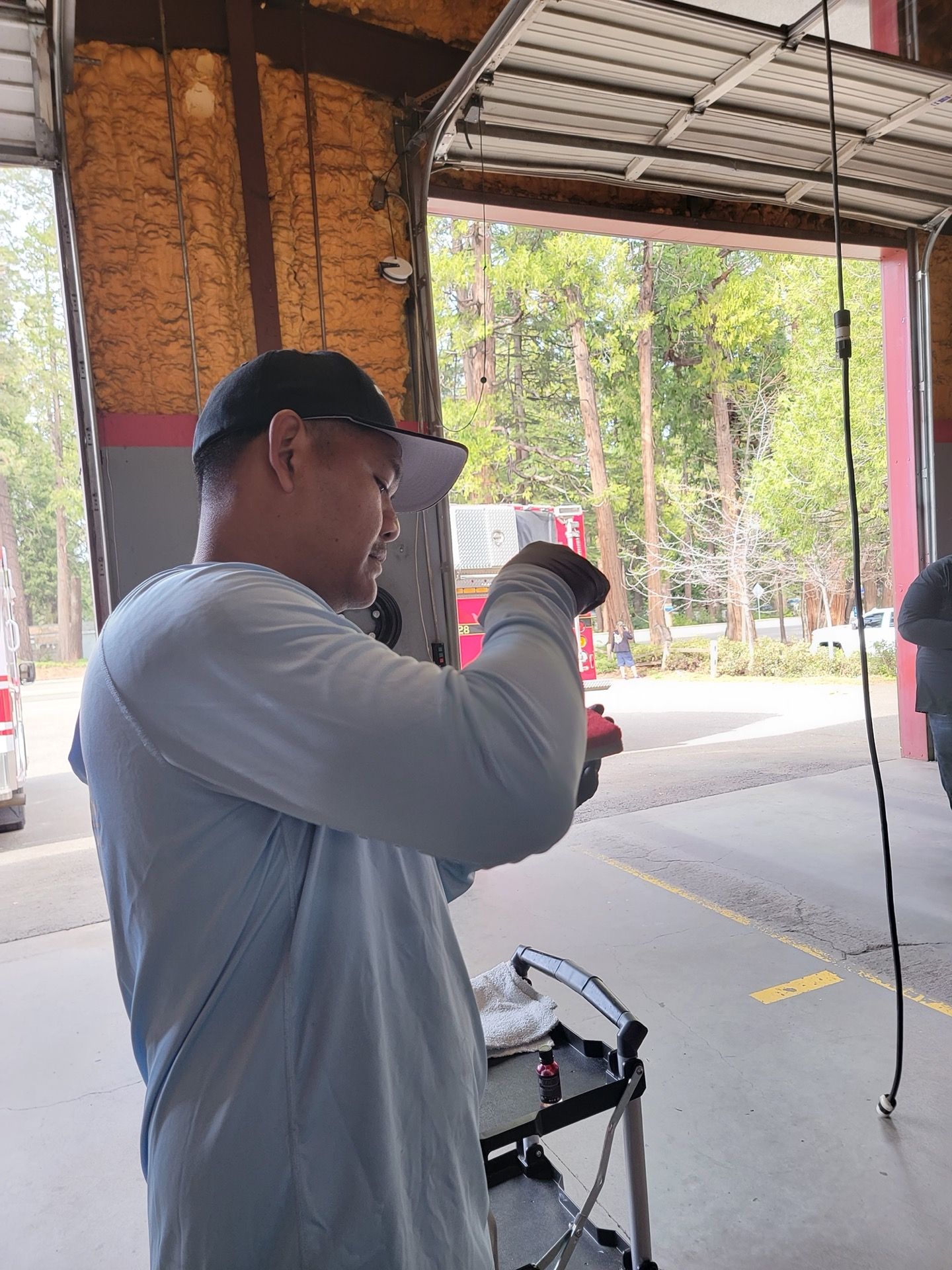 Man in light blue shirt and black cap examines something in an open garage. Outdoors, trees visible.