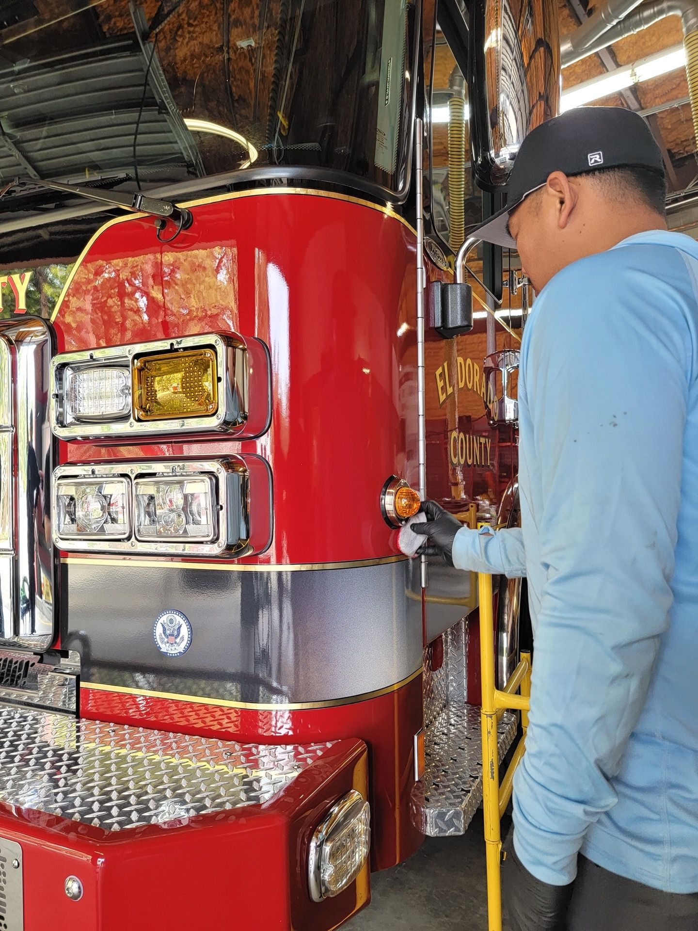 Man in blue shirt cleans the amber light of a red fire truck in a garage.